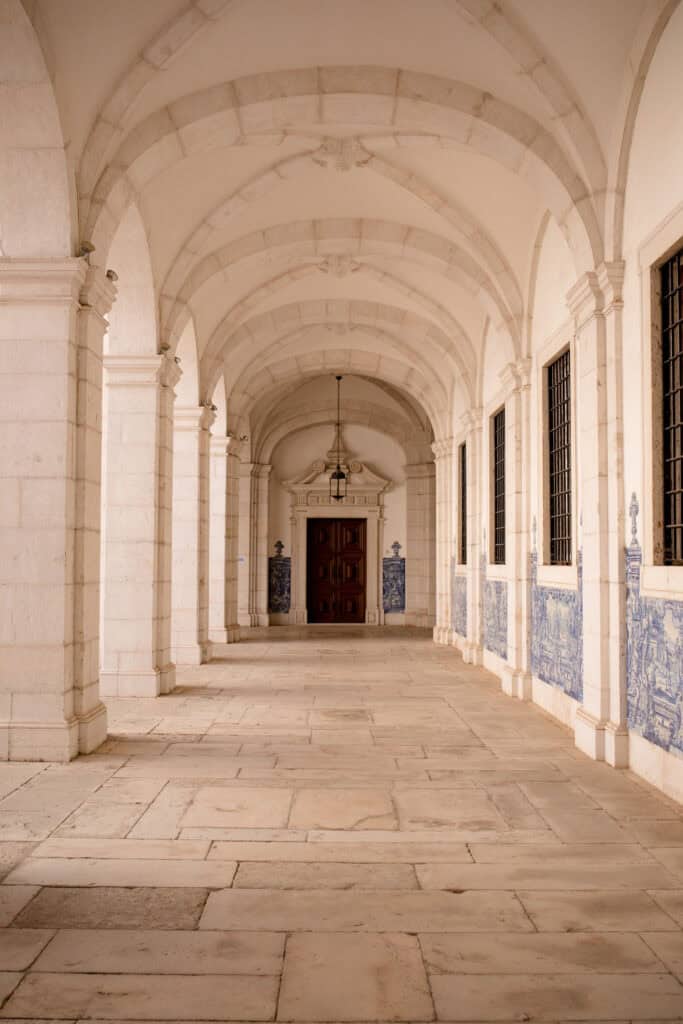 Corredor histórico em Lisboa com arcos de pedra clara e painéis de azulejos azuis e brancos ao longo das paredes. A arquitetura ornamentada e os azulejos tradicionais ajudam a mostrar o tipo de património que pode ser descoberto ao visitar museus em Lisboa.