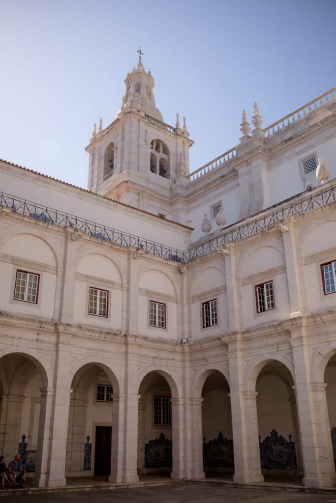 Alt text: Claustro do Mosteiro de São Vicente de Fora em Lisboa com arcadas de pedra branca e painéis de azulejos azuis e brancos nas paredes. A torre da igreja ergue se acima do pátio interior sob um céu azul.