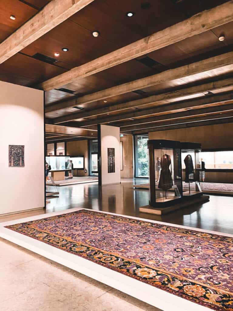 Galeria do Museu Gulbenkian em Lisboa com um grande tapete persa exposto no chão vitrines com peças históricas e visitantes ao fundo. Entre os museus em Lisboa a Gulbenkian destaca se pela coleção de arte apresentada em salas amplas e contemporâneas.