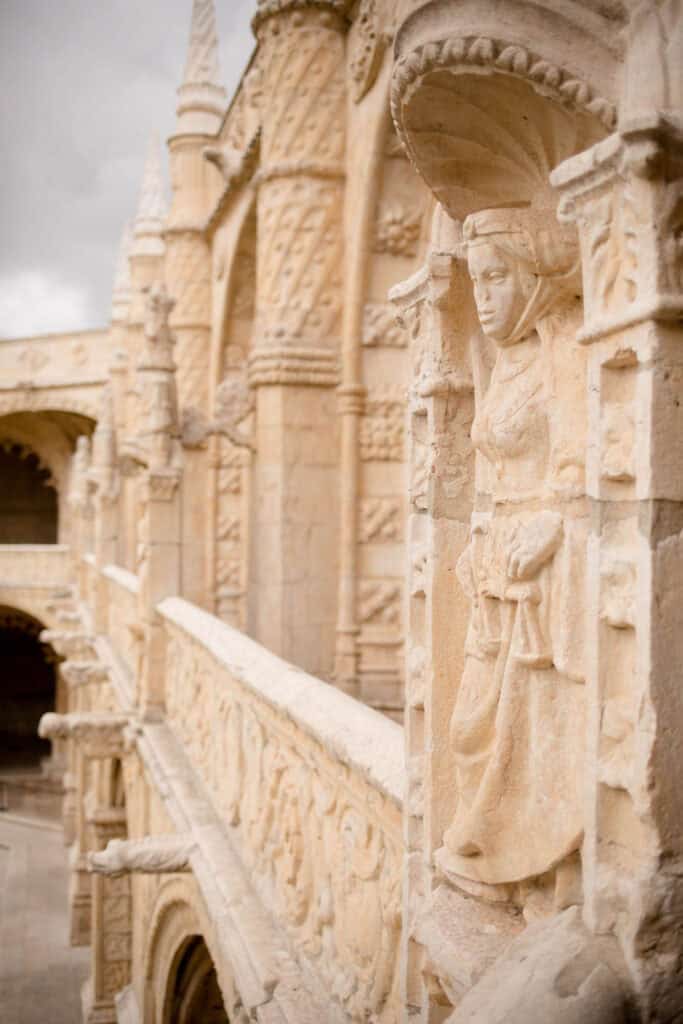 Detalhe em pedra esculpida no claustro do Mosteiro dos Jerónimos em Lisboa com uma figura humana integrada na ornamentação manuelina. Ao fundo aparecem arcos e colunas decoradas em pedra clara que destacam a riqueza arquitetónica do mosteiro dos Jerónimos.