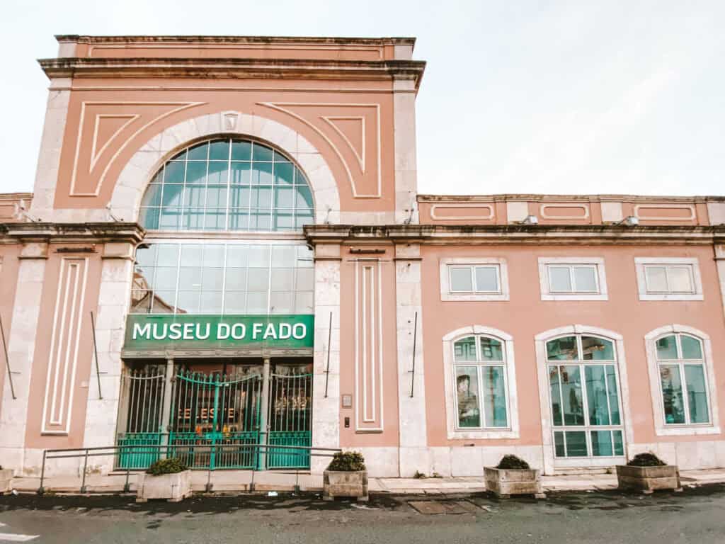 Fachada cor de rosa do Museu do Fado em Lisboa com a entrada marcada pela placa "MUSEU DO FADO" e grandes janelas emolduradas em pedra clara. Visitar o museu é uma das atividades em Lisboa com chuva para conhecer a história e a tradição do fado português.