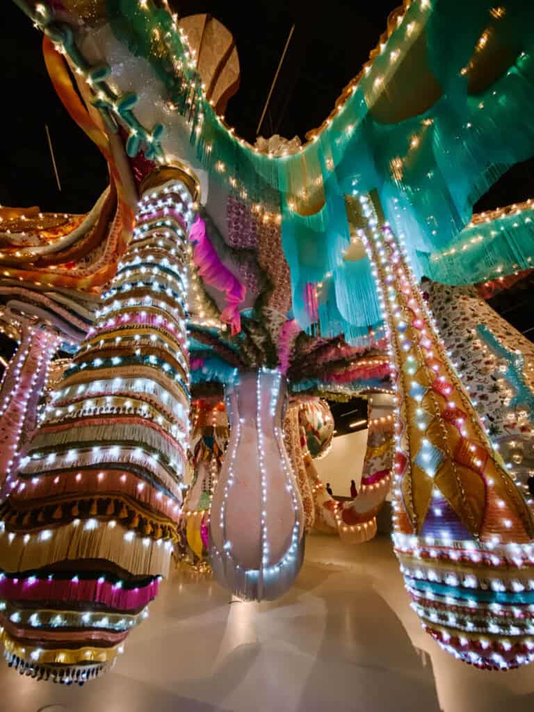 Instalação artística imersiva com formas têxteis gigantes coloridas iluminadas por centenas de pequenas luzes em tons de rosa azul verde e dourado. A exposição mostra o tipo de experiência visual contemporânea que pode ser encontrada em museus em Lisboa, como o MAAT.