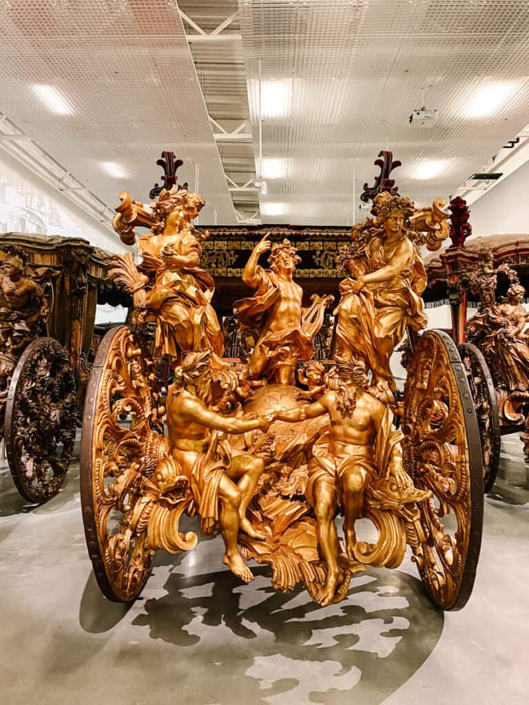 Coche histórico ricamente ornamentado com grandes esculturas douradas de figuras humanas e detalhes barrocos exposto no Museu Nacional dos Coches em Lisboa. A visita ao museu é uma das opções de passeios cobertos em Lisboa para conhecer de perto carruagens reais e arte decorativa.