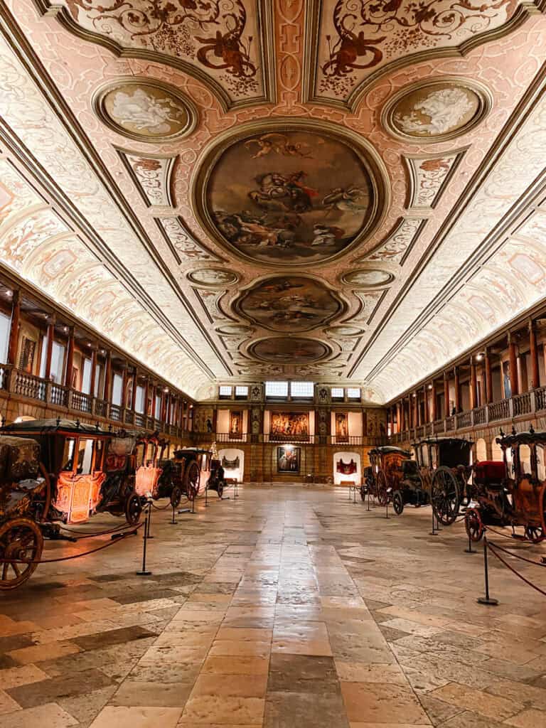 Grande galeria do Museu Nacional dos Coches em Lisboa com carruagens históricas alinhadas dos dois lados e um teto ricamente pintado com painéis decorativos. O salão amplo destaca a coleção de coches reais e a arquitetura histórica do museu nacional dos coches Lisboa.