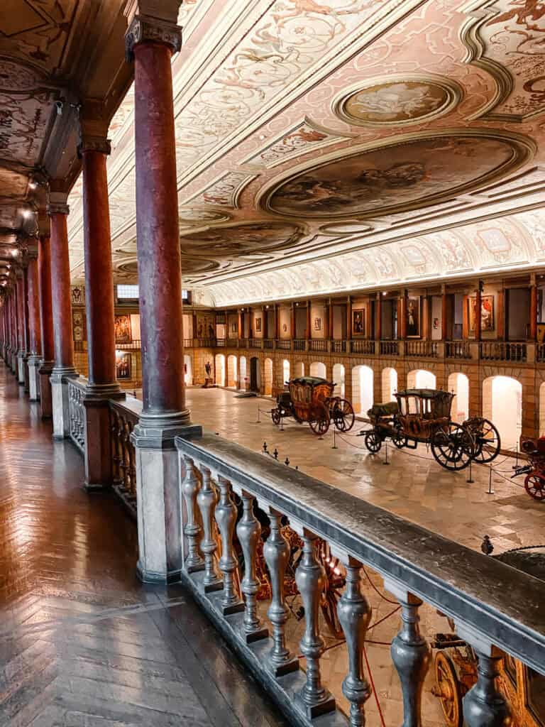 Galeria histórica do Museu dos Coches em Lisboa vista de uma varanda com colunas vermelhas teto pintado e vários coches antigos expostos no piso inferior. O espaço destaca a coleção de carruagens reais e a arquitetura ornamentada do museu dos coches.