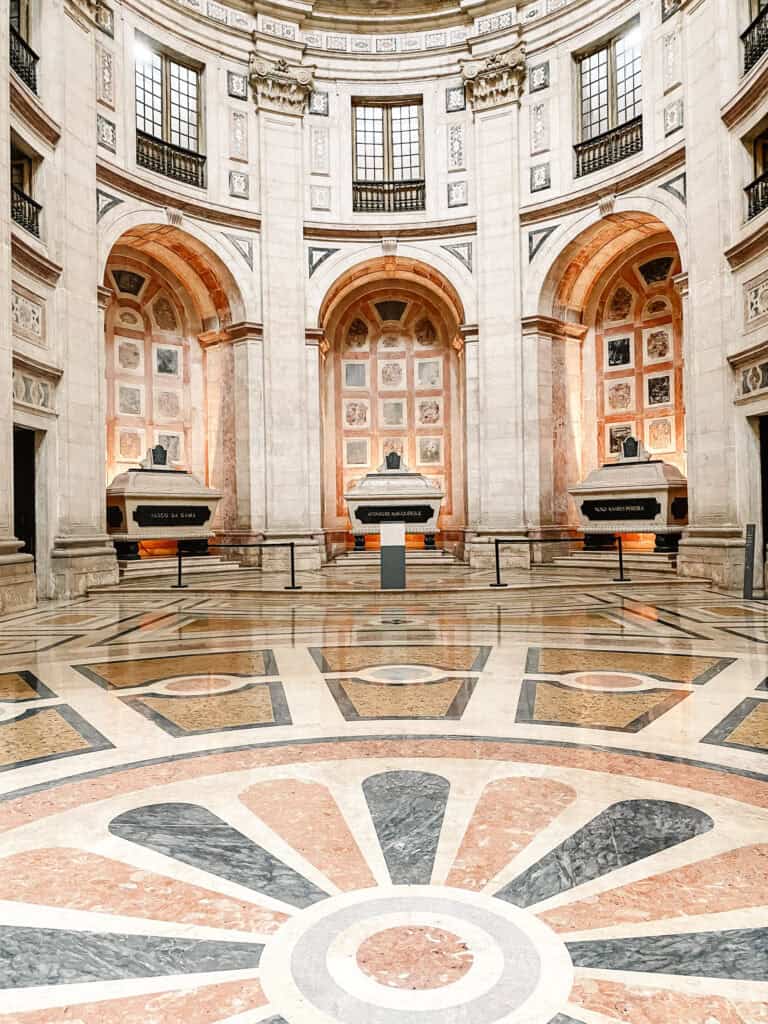 Interior monumental do Panteão Nacional em Lisboa com chão de mármore geométrico e três túmulos sob grandes arcos de pedra. As inscrições dizem "VASCO DA GAMA" "AFONSO DE ALBUQUERQUE" e "NUNO ÁLVARES PEREIRA" num espaço histórico coberto que é uma opção para quem procura o que fazer em Lisboa com chuva.