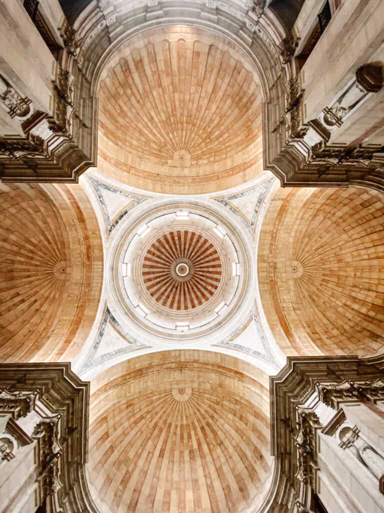 Vista para cima da cúpula do Panteão Nacional em Lisboa com padrões radiais em pedra clara e tons avermelhados cercados por grandes arcos ornamentados. O interior coberto do monumento mostra uma das atividades em Lisboa com chuva para quem quer explorar arquitetura e história.