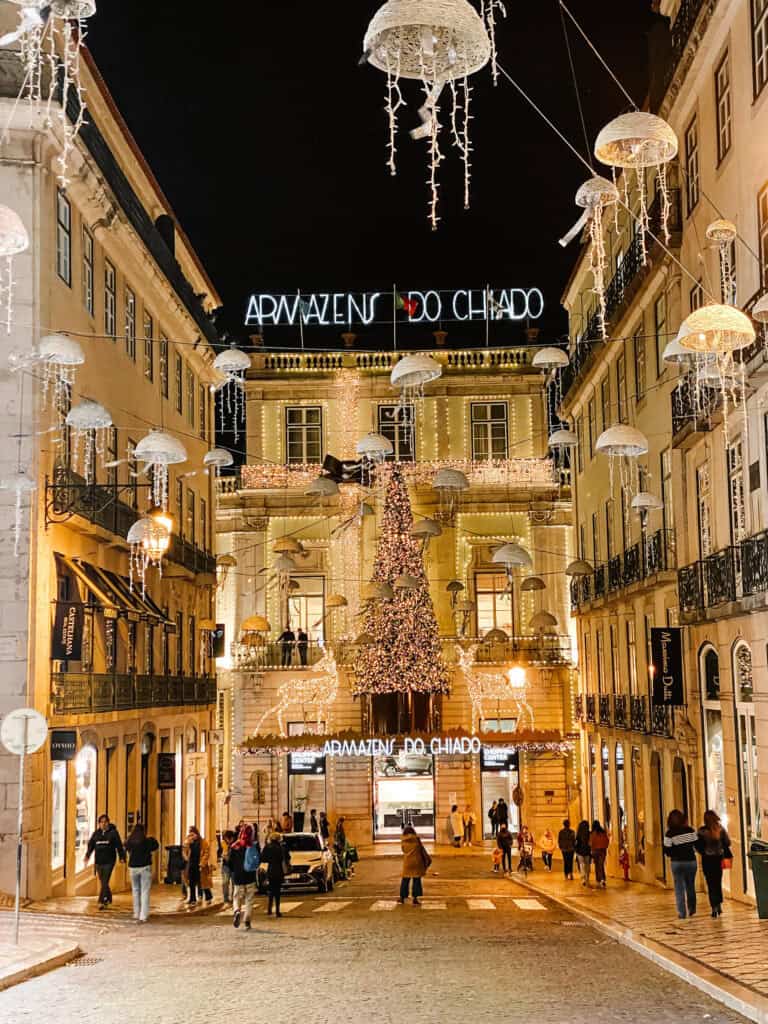 Rua do Chiado iluminada à noite com decorações de Natal suspensas e uma grande árvore iluminada diante do "ARMAZÉNS DO CHIADO". A zona comercial cheia de lojas mostra uma opção popular para fazer compras em Lisboa no inverno.