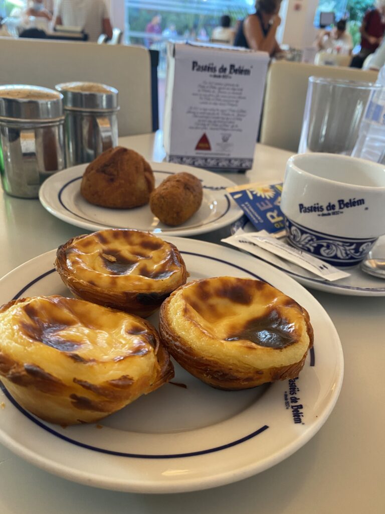 Três pastéis de nata servidos num prato na pastelaria "Pastéis de Belém" com uma chávena da casa e outros salgados ao fundo. Uma paragem coberta para provar os famosos pastéis é uma opção agradável para explorar Lisboa com crianças em dias de chuva.