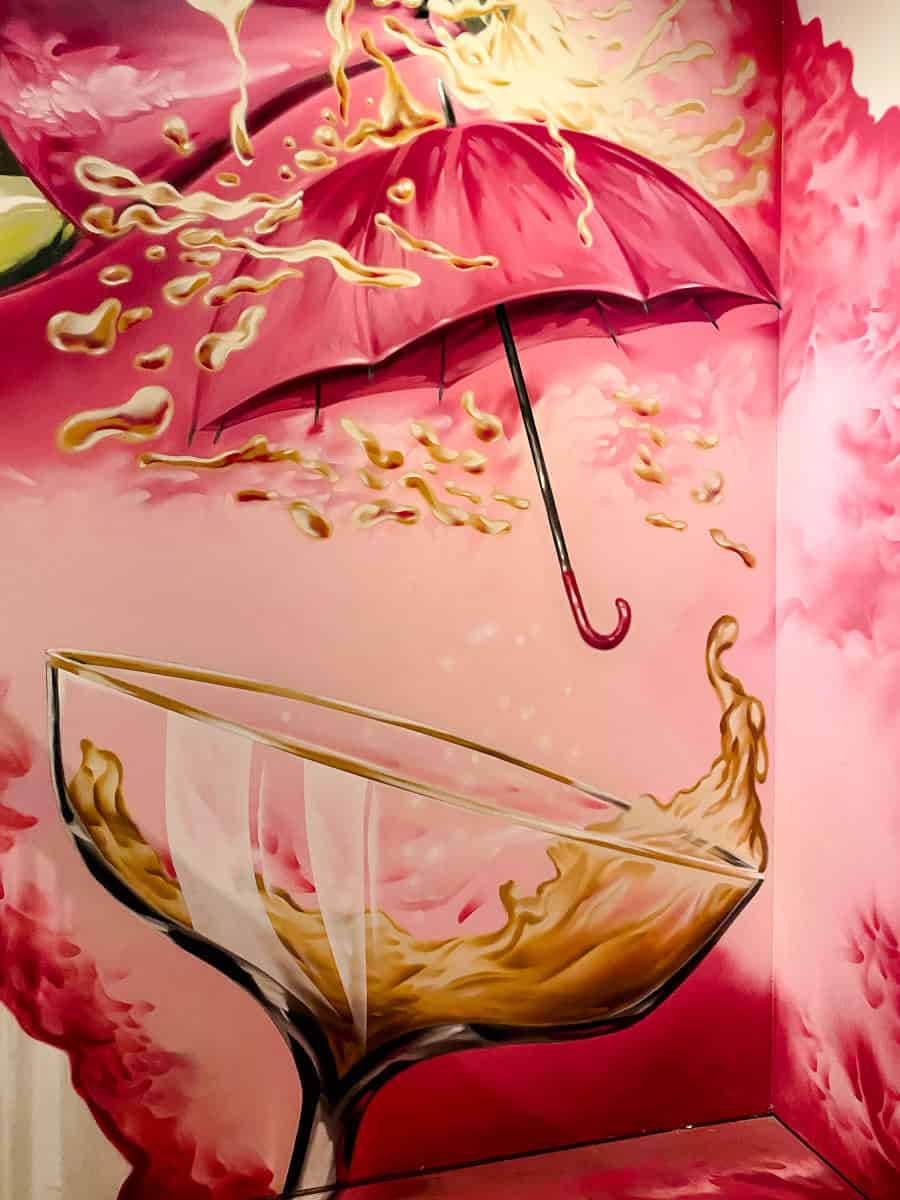 Een levendige muurschildering van een champagnecoupe die overloopt met bubbels die tegen een felroze paraplu spatten in de Pink Palace WOW.
