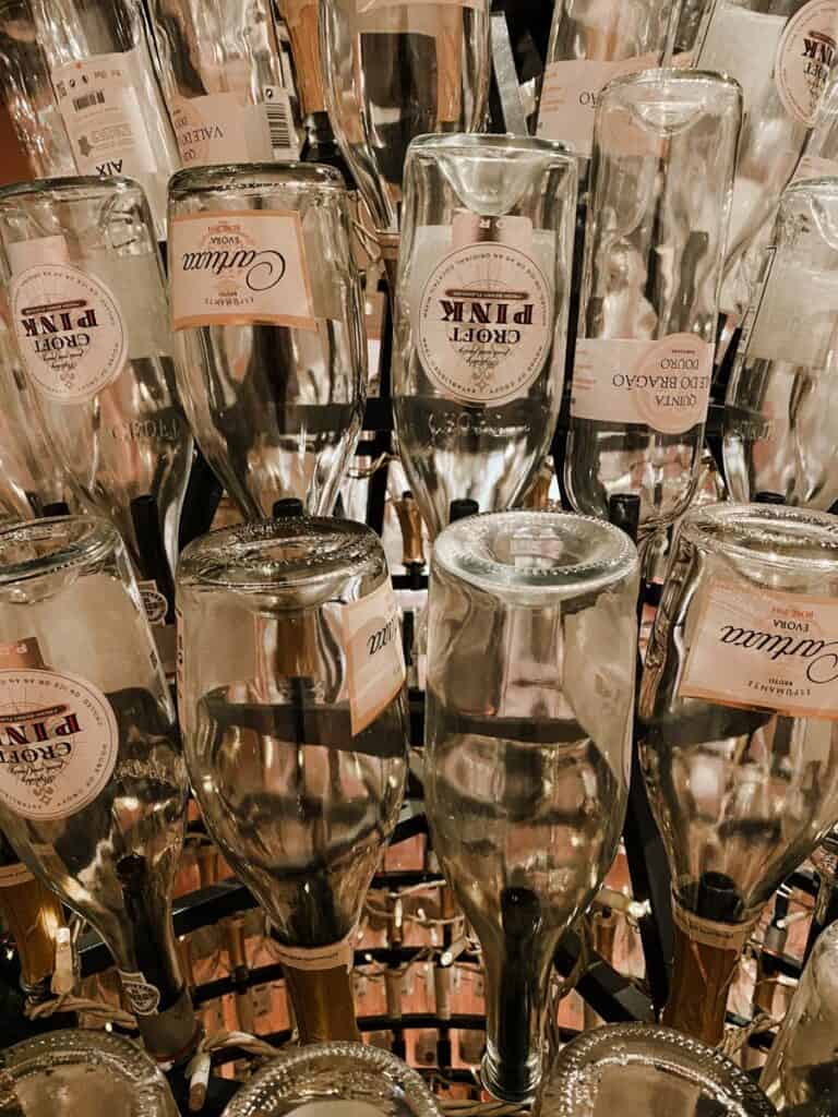 Garrafas de vinho rosé penduradas de cabeça para baixo em uma estrutura circular com iluminação quente ao fundo, criando um efeito decorativo elegante no Pink Palace Porto. Rótulos visíveis como “Croft Pink” e “Espumante Aurora Rosé” aparecem nas garrafas organizadas em padrão repetido.