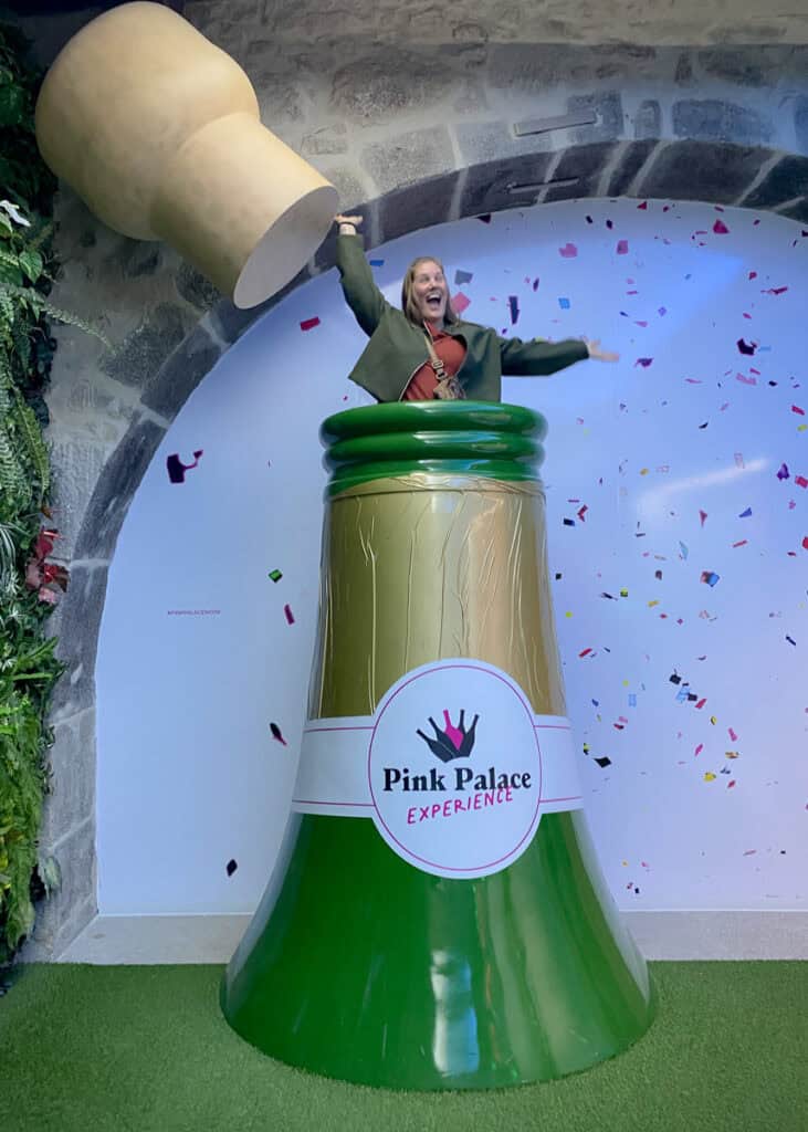 Een vrouw springt lachend uit een gigantische champagnefles met confetti in de lucht tijdens de Pink Palace experience.