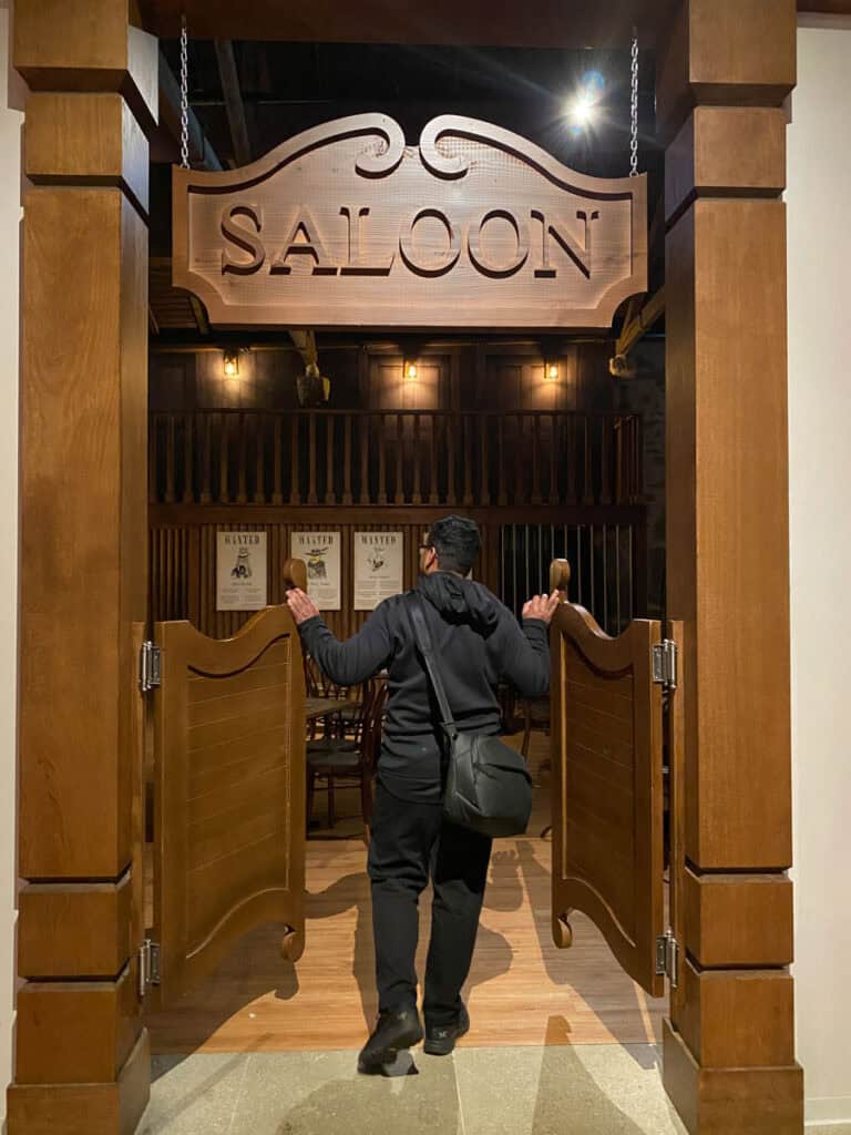 Entrada temática de saloon com portas estilo faroeste e placa de madeira escrita “SALOON”, enquanto uma pessoa entra no ambiente decorado com iluminação quente no Pink Palace Porto. A cena mostra uma das áreas interativas incluídas na experiência, comum em conteúdos sobre pink palace porto ingressos.