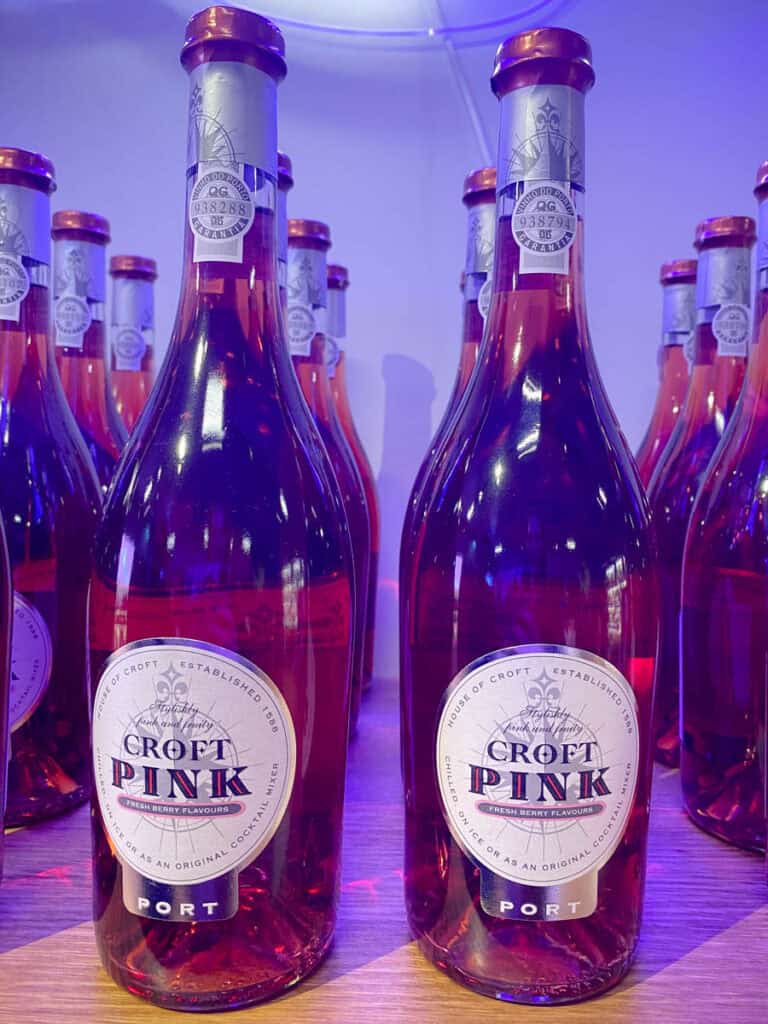 Garrafas de vinho rosé Croft Pink alinhadas sobre uma superfície com iluminação em tons de roxo e rosa, destacando os rótulos “Croft Pink” e “Port”. A cena representa a exposição de vinhos no museu do rosé Porto com foco na estética vibrante da experiência.
