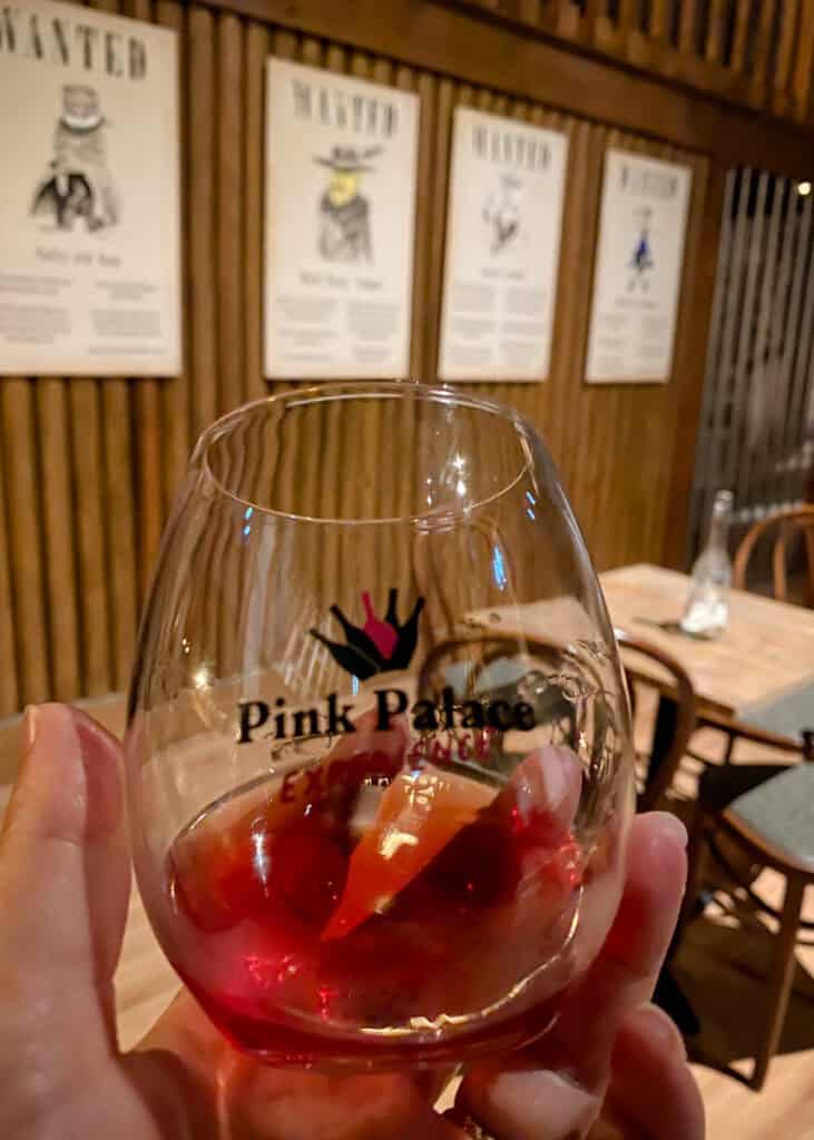Mão segurando uma taça com vinho rosé e o texto “Pink Palace Experience”, com mesas de madeira e cartazes estilo “WANTED” ao fundo em um ambiente temático no WOW Porto em Vila Nova de Gaia. A cena destaca a degustação em um espaço decorado que combina experiência interativa e atmosfera de bar.