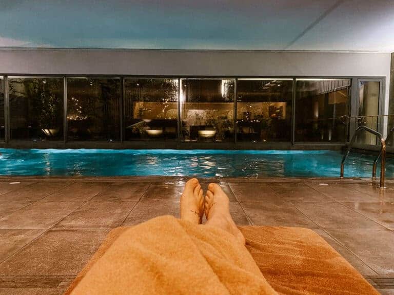 Pessoa relaxa numa espreguiçadeira diante de uma piscina interior iluminada em tons de azul num ambiente de spa. Passar algumas horas num spa com piscina aquecida é uma opção tranquila de passeios cobertos em Lisboa para escapar à chuva.