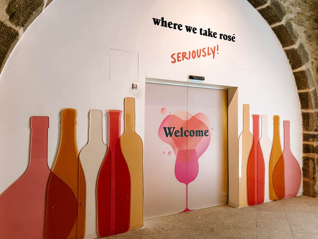 Entrada com portas decoradas com ilustração de taça rosa e a palavra “Welcome”, cercada por desenhos de garrafas em tons de rosé e a frase “where we take rosé SERIOUSLY!” na parede. O espaço marca a chegada ao Pink Palace no WOW Porto, atração popular para quem busca o que fazer no WOW Porto.