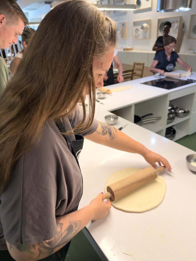 Mulher estende massa com um rolo durante uma aula de pastel de nata numa cozinha de confeitaria em Lisboa enquanto outros participantes trabalham ao fundo. A experiência prática é uma das atividades em Lisboa com chuva para aprender a preparar um dos doces mais tradicionais da cidade.