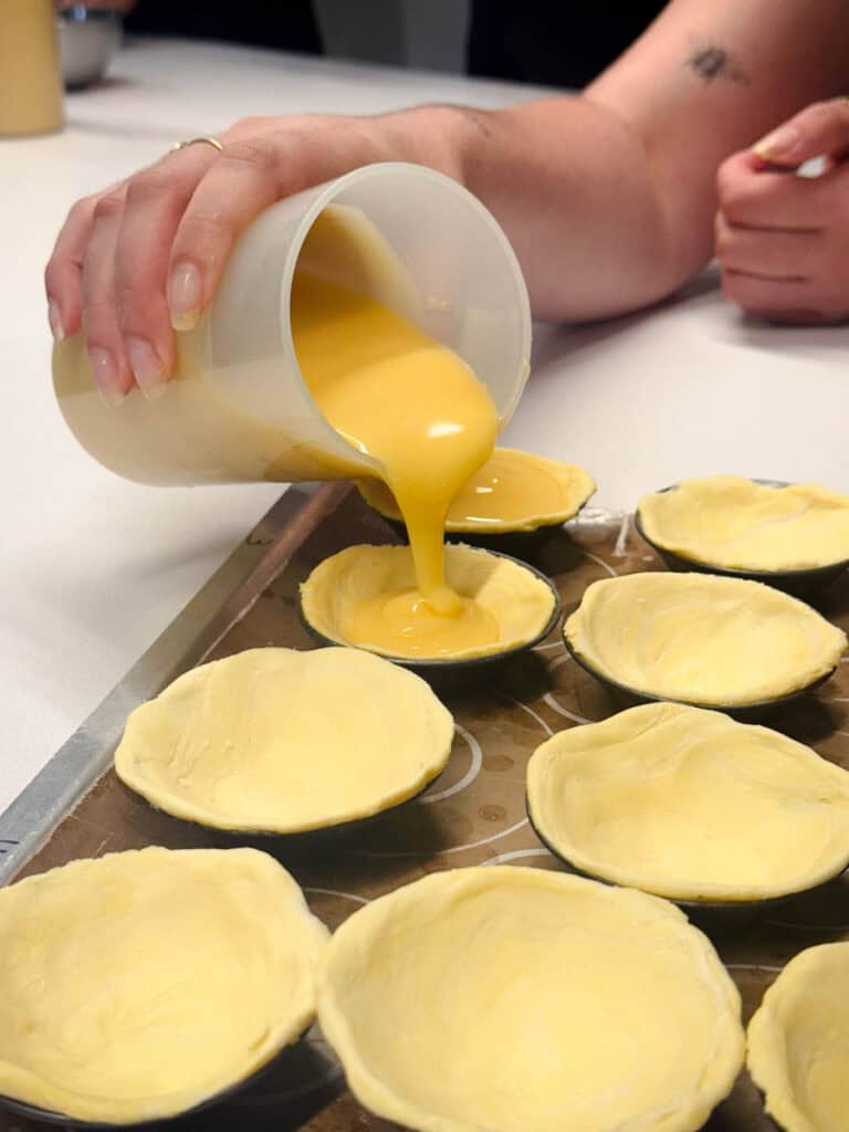 Pessoa despeja creme amarelo em pequenas formas forradas com massa durante uma aula de pastel de nata em Lisboa. A experiência culinária em espaço fechado é uma opção acolhedora para aproveitar Lisboa no inverno.