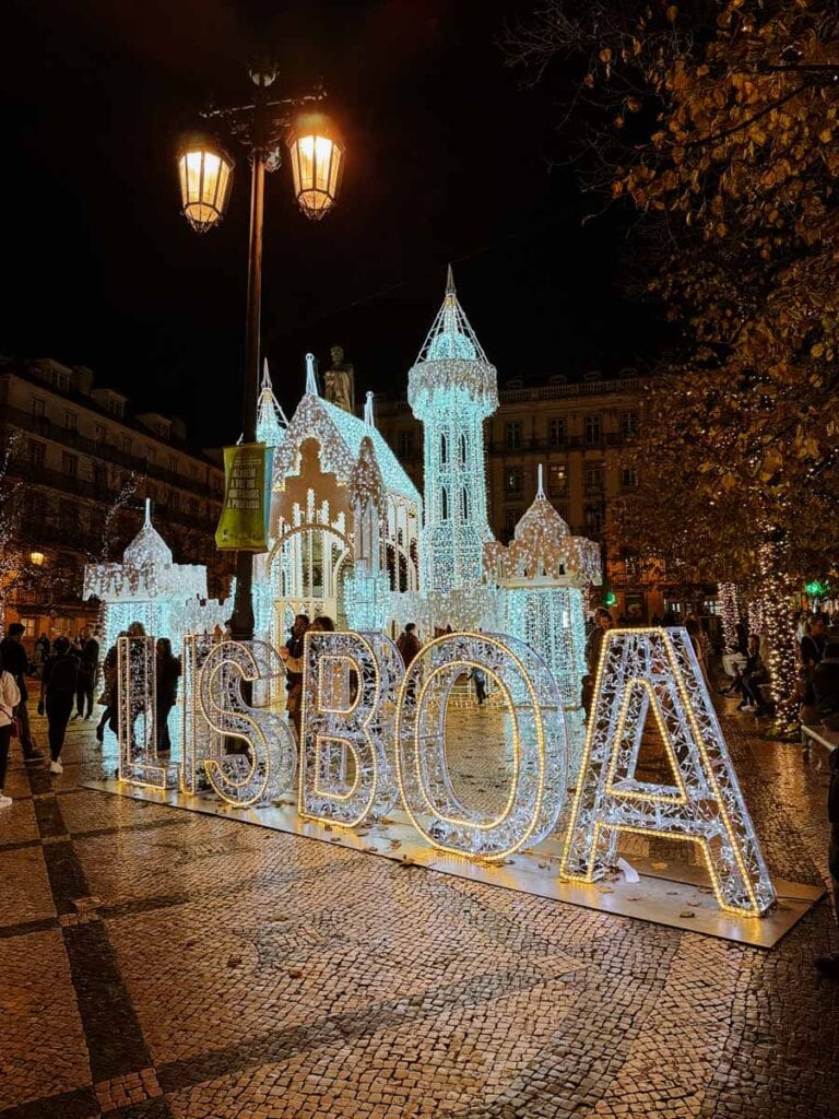 An illuminated fairytale castle stands behind giant glowing “LISBOA” letters in a Lisbon square, with trees and buildings adorned in twinkling lights. This magical scene is one of the most whimsical Lisbon Christmas lights installations and a festive highlight among Christmas decorations in Lisbon.