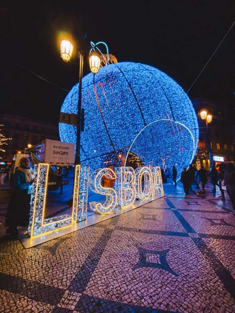 A towering blue light-up bauble dominates the scene behind the illuminated “LISBOA” sign, glowing brightly in a busy square under warm streetlamps. This bold and immersive setup is a major attraction of the Lisbon Christmas lights and one of the most photographed Christmas lights in Lisbon.