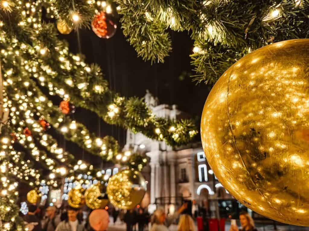 Close-up of golden baubles and glowing string lights on a Christmas tree in Lisbon’s Praça do Comércio, with the Arco da Rua Augusta softly lit in the background. The festive scene captures the sparkling detail of Christmas lights Lisbon is known for.