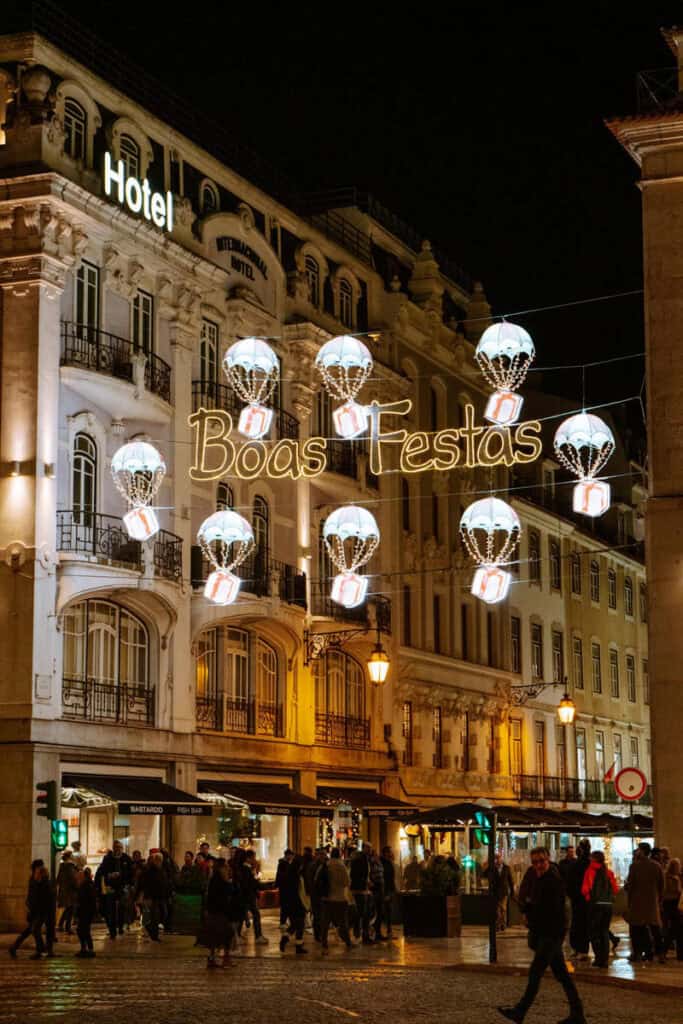 Hot-air balloon style lights carrying presents float above a Lisbon street, surrounding the glowing “Boas Festas” sign strung between two historic buildings. The display is a playful and welcoming part of Lisbon Christmas lights and showcases the creativity of Christmas decorations in Lisbon.