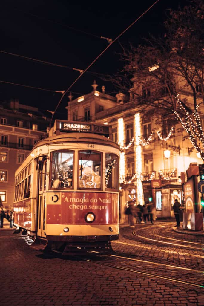 A classic red tram on route 28 rolls through Lisbon at night, decorated for the season with the words “A magia do Natal chega sempre” and surrounded by fairy-lit buildings. The festive tram adds charm to Lisbon Christmas lights and showcases how Christmas decorations in Lisbon extend to its iconic transport.