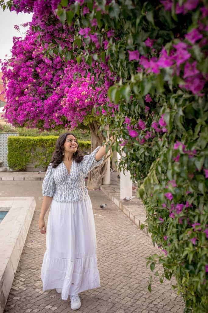 Een vrouw in een witte rok en luchtige blouse wandelt glimlachend langs een muur vol felroze bougainville in Alfama. De ontspannen sfeer en kleurrijke bloemen maken deze Alfama fotoshoot levendig en zomers.