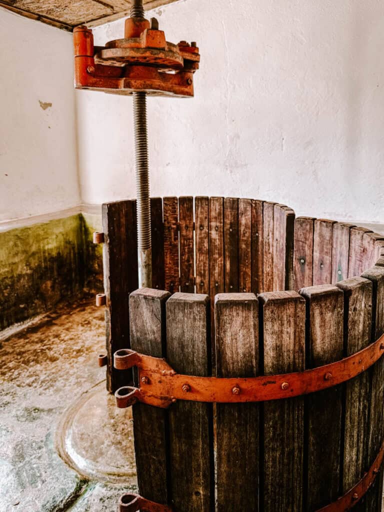 An old wooden and metal wine press inside a whitewashed room, showing signs of use and age. Highlights traditional winemaking methods still appreciated on a Lisbon vineyard tour.