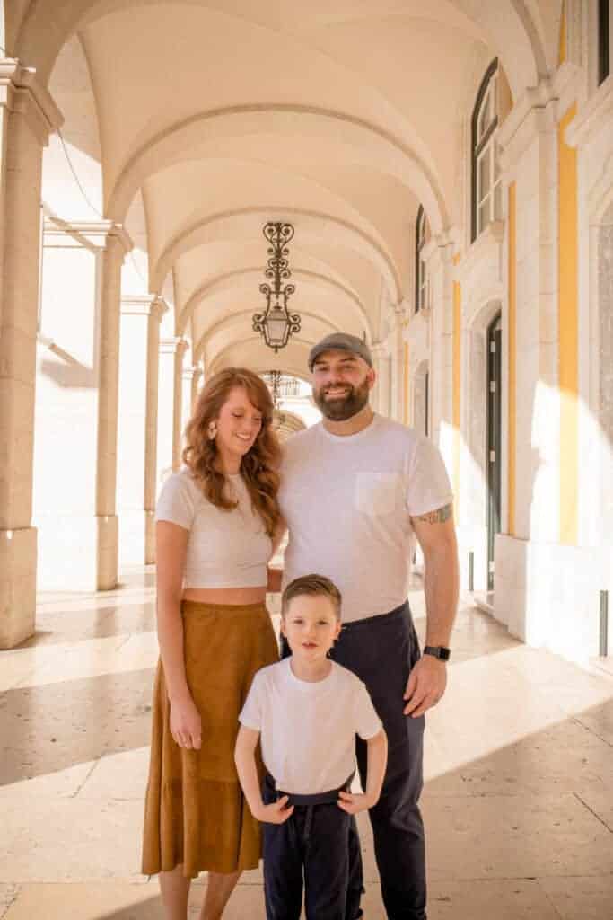 Een gezin van drie poseert onder een met bogen versierde galerij in Lissabon, badend in warm zonlicht. De moeder, vader en zoon dragen op elkaar afgestemde, lichte outfits, wat zorgt voor een harmonieuze uitstraling passend bij familie fotografie in Lissabon.