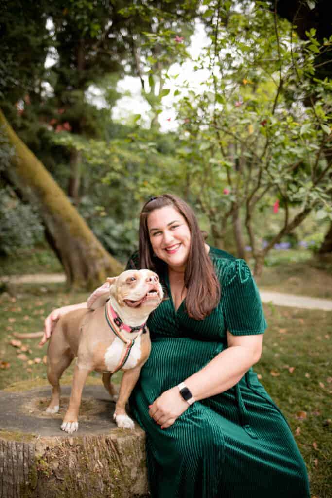 Een lachende vrouw in een donkergroene fluwelen jurk poseert met haar beige pitbull op een boomstronk in een groen park in Lissabon. De ontspannen en natuurlijke sfeer benadrukt de stijl van een fotograaf in Lissabon.