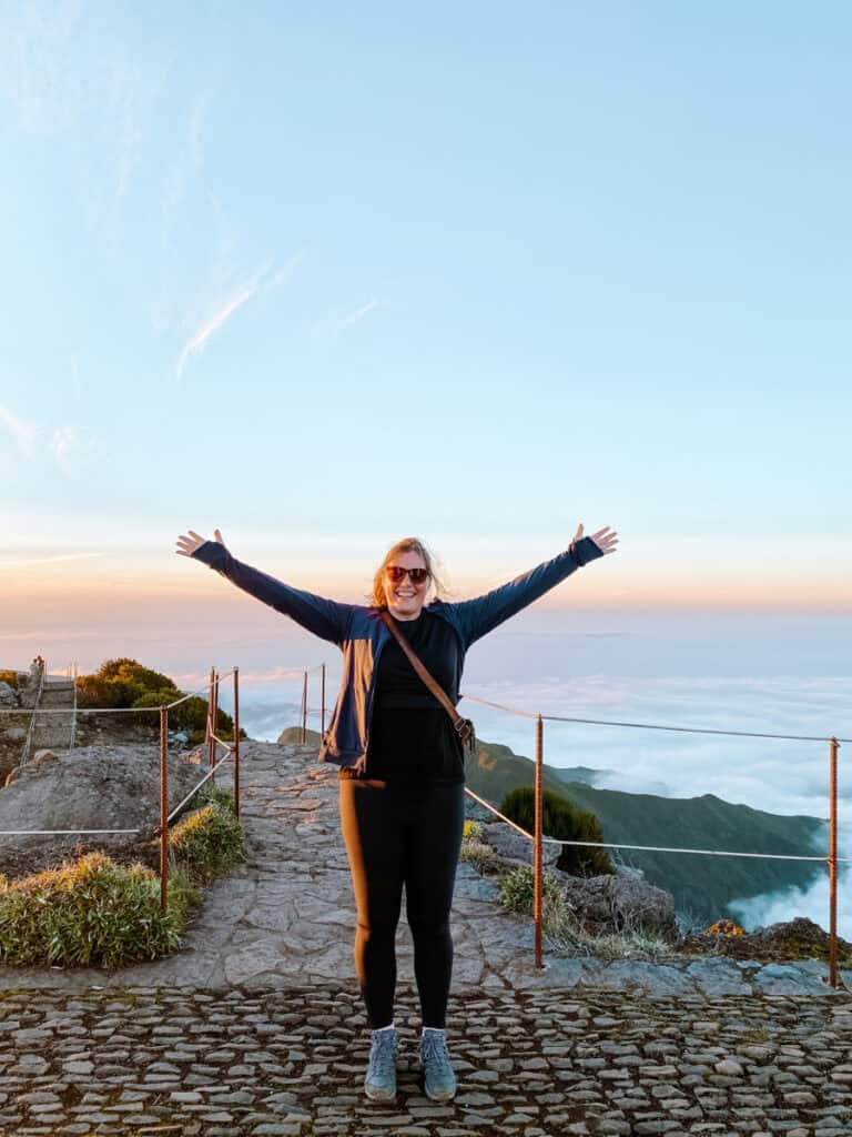 Wandelaar op de Pico Ruivo hike die glimlachend over een geplaveid pad met metalen leuningen loopt, vlak voor de top. Het warme licht van de zonsondergang schijnt over de ruige bergroute.