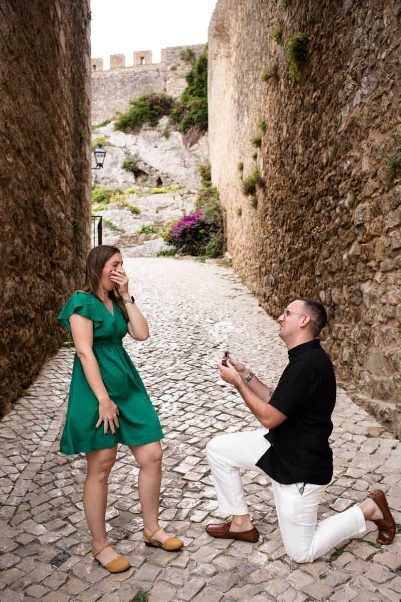 Een man gaat op één knie voor zijn partner in een smal, historisch steegje in Óbidos, omringd door stenen muren en bloemen. Haar emotionele reactie en zijn ringdoosje leggen het magische moment van dit huwelijksaanzoek in Óbidos perfect vast.