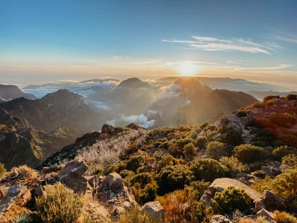 Zonsondergang bij Pico Ruivo op Madeira met gouden zonlicht dat over de ruige bergtoppen en wolkenzee schijnt. Op de voorgrond zie je rotsen en lage struiken langs de Vereda do Pico Ruivo hike.