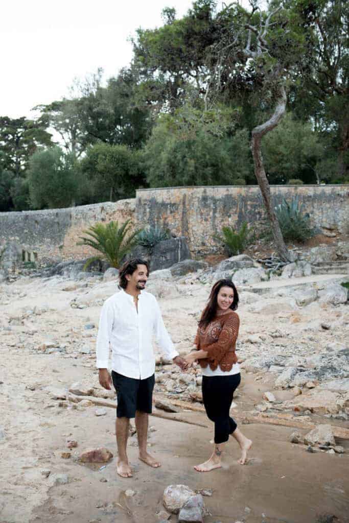 Een stel loopt hand in hand op blote voeten langs een rotsachtig strand in Cascais, omringd door bomen en een verweerde stenen muur. Hun gelukkige blikken en ontspannen houding stralen liefde uit, perfect voor een verlovingsfotoshoot in Cascais.
