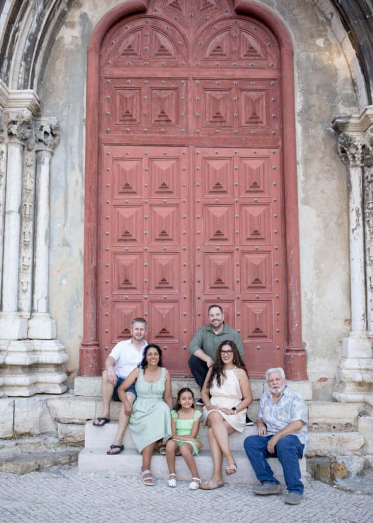 Multigenerational family of six seated on stone steps in front of a grand red wooden church door in Bairro Alto, Lisbon. The group smiles at the camera, framed by ornate stone columns and historic architecture that highlights the character of the neighbourhood. Relaxed outdoor portrait from a family photo session in Bairro Alto capturing connection across generations in the heart of the city.
