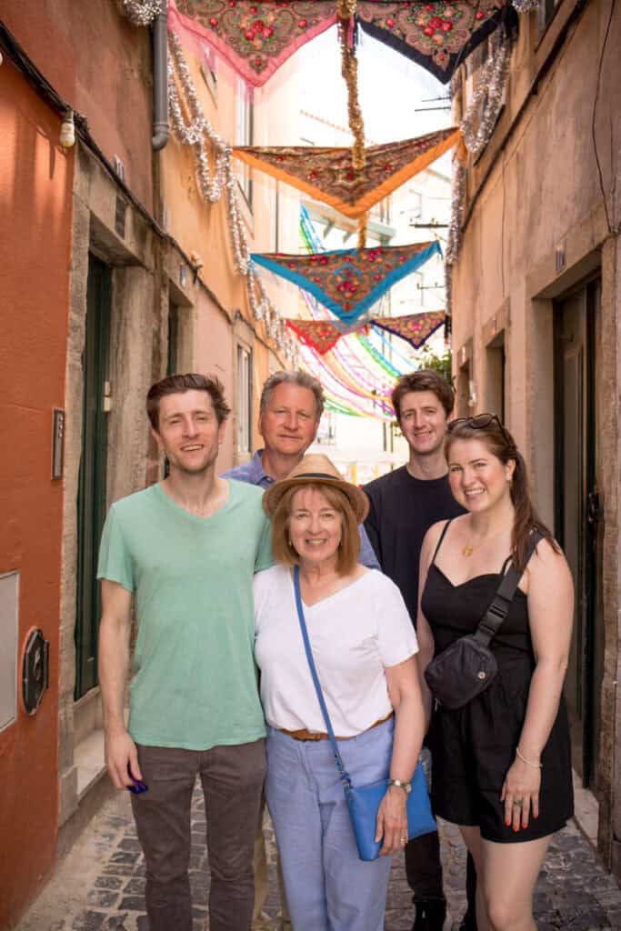 Family of five standing together in a narrow Lisbon street decorated with colourful festival garlands overhead. The group smiles at the camera, framed by pastel buildings and cobbled paving that add character to the scene. Relaxed portrait from a family photo shoot Lisbon capturing togetherness and the vibrant atmosphere of the city.