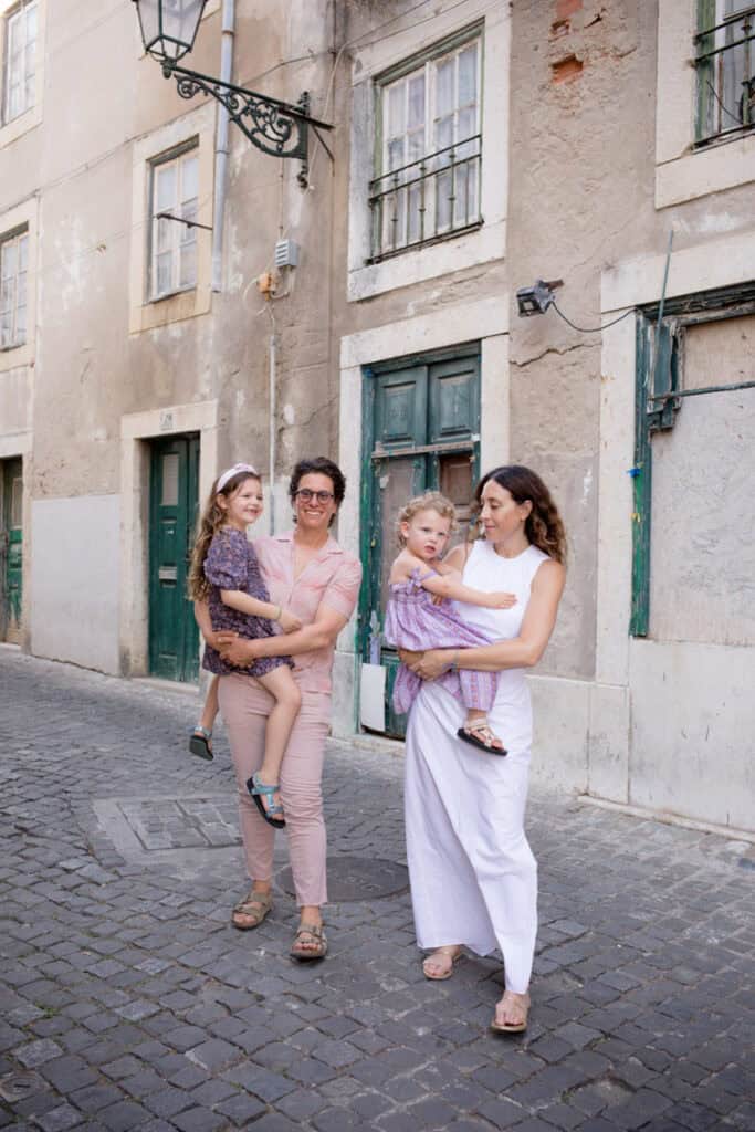 Two mothers walking along a cobbled street in Lisbon, each holding their young daughter in their arms against a backdrop of pastel buildings and traditional green doors. The women smile at one another while the girls in patterned dresses look around, capturing a relaxed and affectionate moment. Natural lifestyle image from a family photoshoot in Lisbon highlighting connection and the charm of the historic neighbourhood.