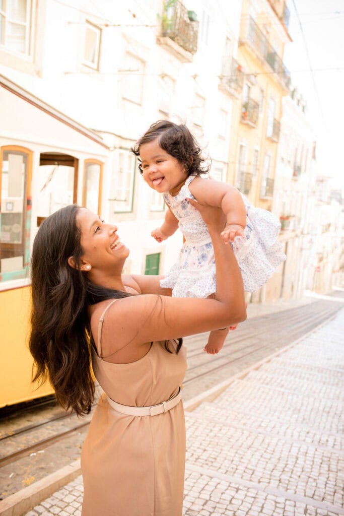 Mother lifting her smiling toddler into the air on a sunlit cobbled street in Lisbon, with a yellow tram and pastel buildings in the background. The little girl in a light patterned dress laughs as they share a playful moment during an outdoor session with a Lisbon family photographer. Natural lifestyle portrait capturing joy and connection in the heart of the city.