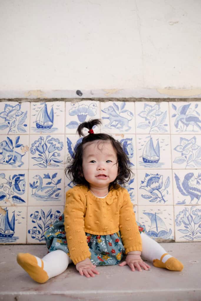 Baby girl sitting on a stone step in front of traditional blue and white azulejo tiles in Lisbon, looking curiously at the camera. She wears a mustard cardigan, floral skirt and matching tights, with her hair tied in a small top knot. Sweet candid portrait from a Lisbon family photoshoot capturing personality against an iconic tiled backdrop.