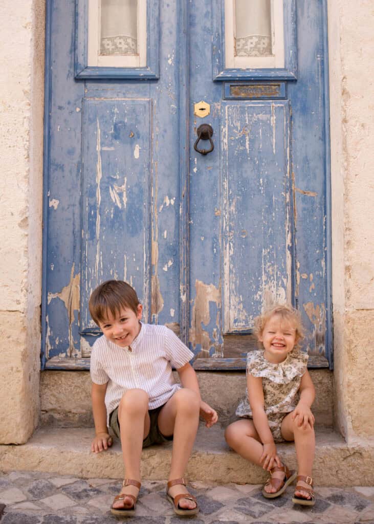 Two young children sitting on stone steps in front of a weathered blue wooden door in Lisbon, smiling brightly at the camera. The boy in a striped shirt and shorts and the little girl in a floral romper wear matching brown sandals, framed by textured walls and cobbled paving. Candid sibling portrait from a Lisbon family photoshoot capturing playful expressions and the charm of the city streets.