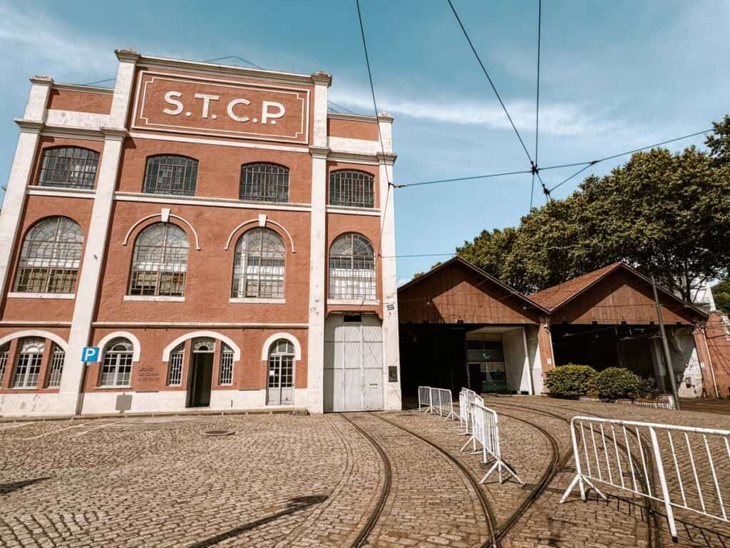 The reddish brown façade of Museu do Carro Eléctrico with tall arched windows and the letters "S.T.C.P" displayed above. Tram tracks cross the cobbled forecourt towards the wooden depot beneath overhead power lines.
