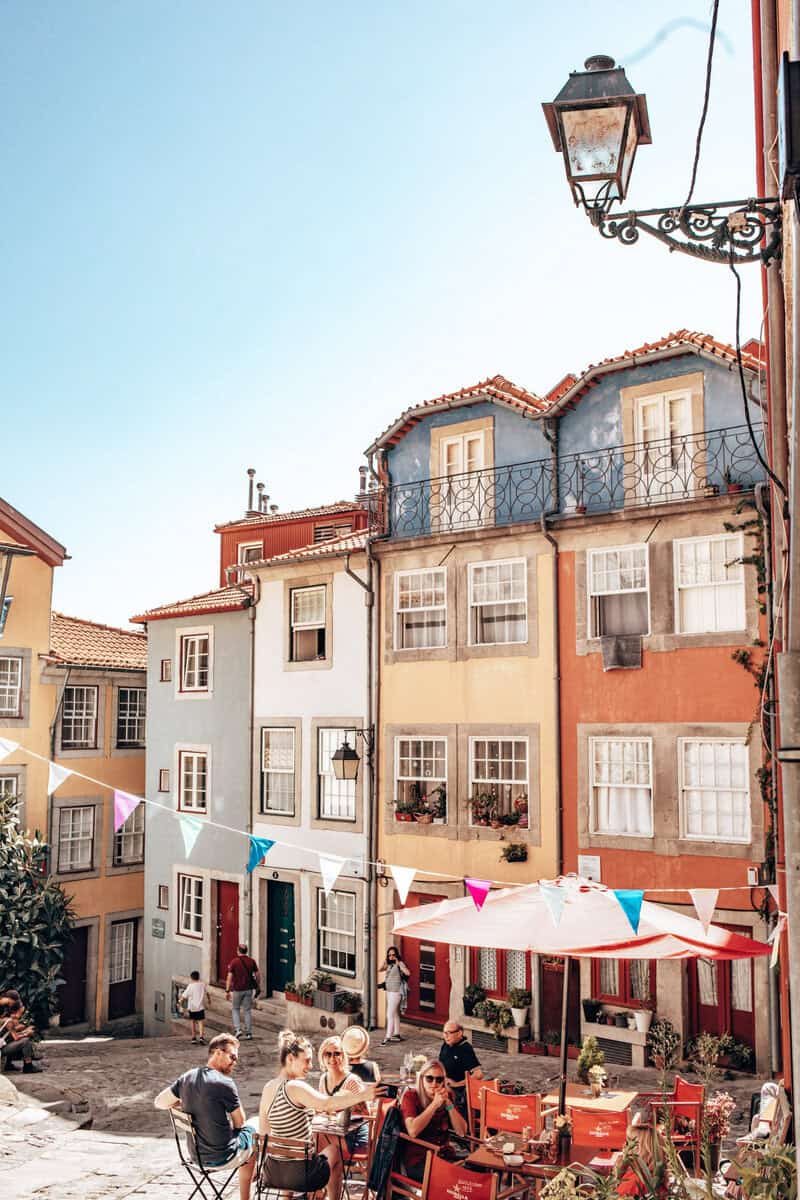 Gezellig pleintje in Porto met kleurrijke huizen op de achtergrond en een terras met rode stoelen en vlaggetjes boven de tafels. Dit soort verborgen plekjes tussen de smalle straatjes zijn ideale Porto vakantie tips voor wie de authentieke sfeer van de stad wil ervaren.