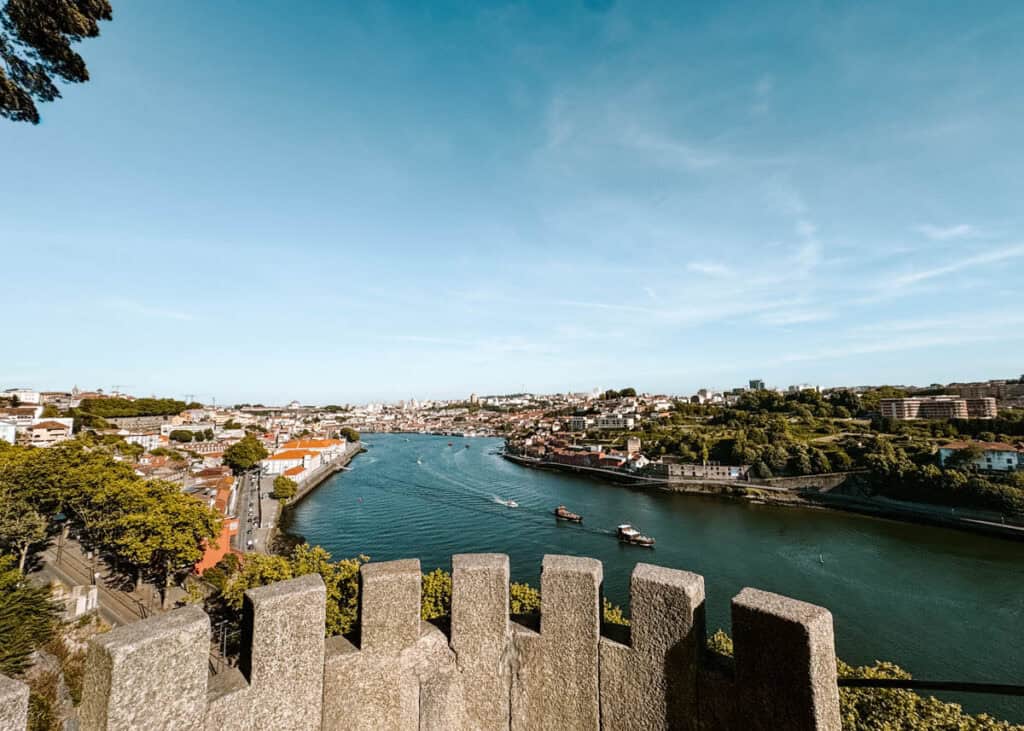 View from Torreão do Jardim do Palácio across the Douro River and the hills of Porto. Stone battlements frame the foreground while boats travel between green riverbanks and terracotta rooftops.