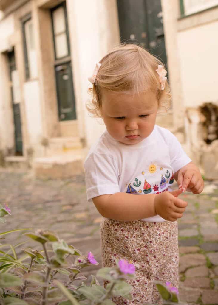 Toddler girl standing on a cobbled Lisbon street, gently touching pink flowers as she explores her surroundings. She wears a white T shirt with colourful sailboat and sun illustrations and floral trousers, with soft curls clipped back by pale bows. Natural candid portrait captured by a family photographer in Lisbon highlighting curiosity and the charm of the old town streets.