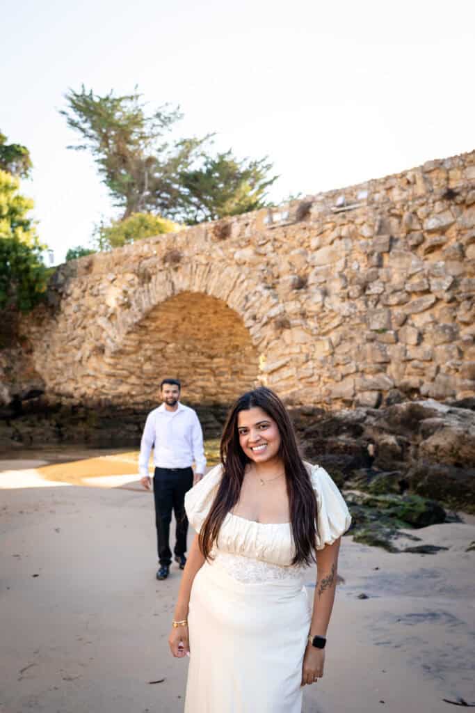 Een lachende vrouw op het strand met haar partner op de achtergrond bij een stenen boog, vastgelegd door een fotograaf voor een aanzoek in Lissabon.