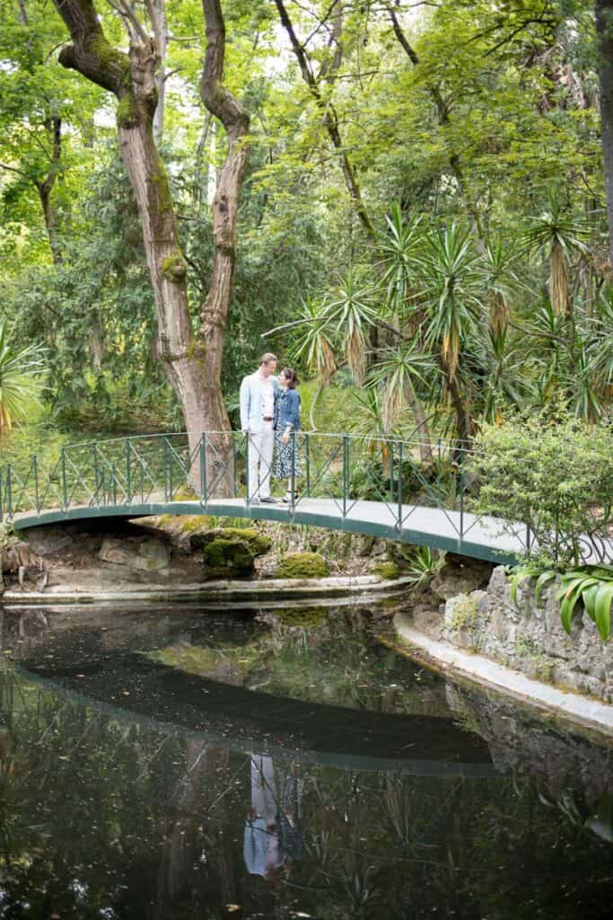 Een romantisch moment tijdens een Lissabon aanzoek waarbij een koppel op een groen bruggetje over een vijver staat, omringd door weelderige bomen en tropische planten.