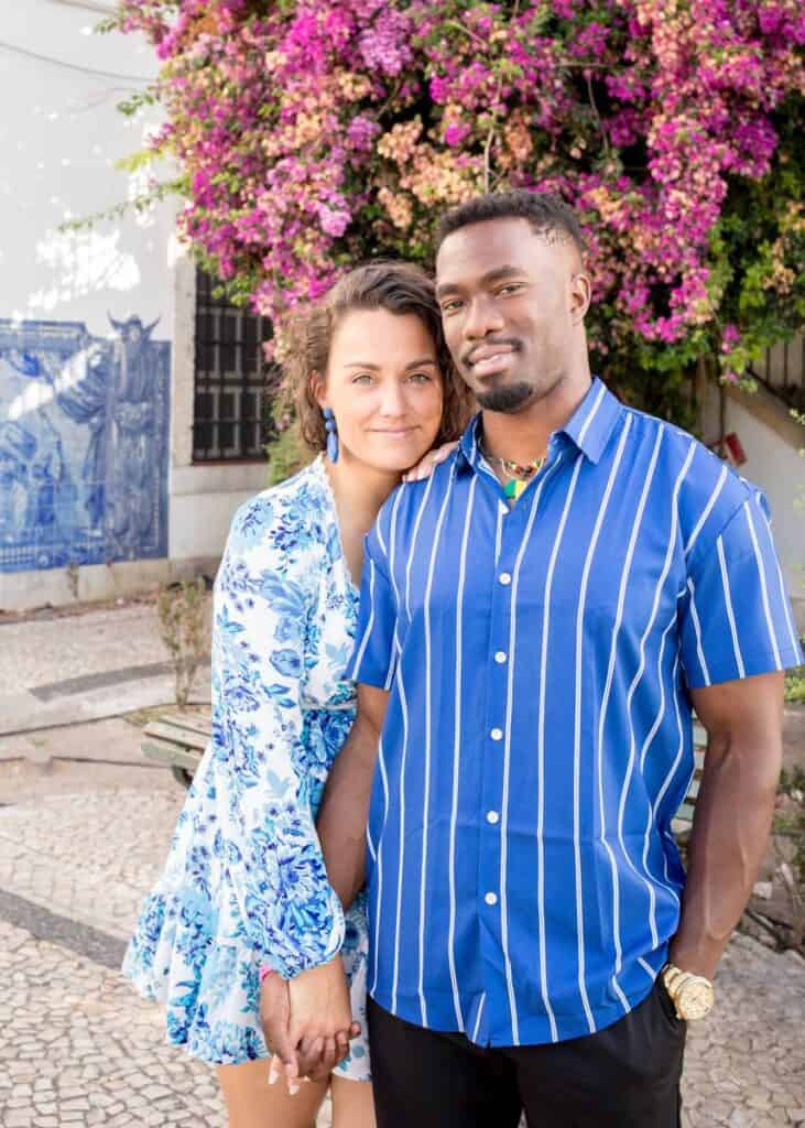 Een gelukkig koppel poseert hand in hand voor hun Lissabon aanzoek bij Miradouro de Santa Luzia, met een achtergrond van levendige paarse bougainvillea en traditionele blauw-witte azulejos (tegels) onder stralend zonlicht.