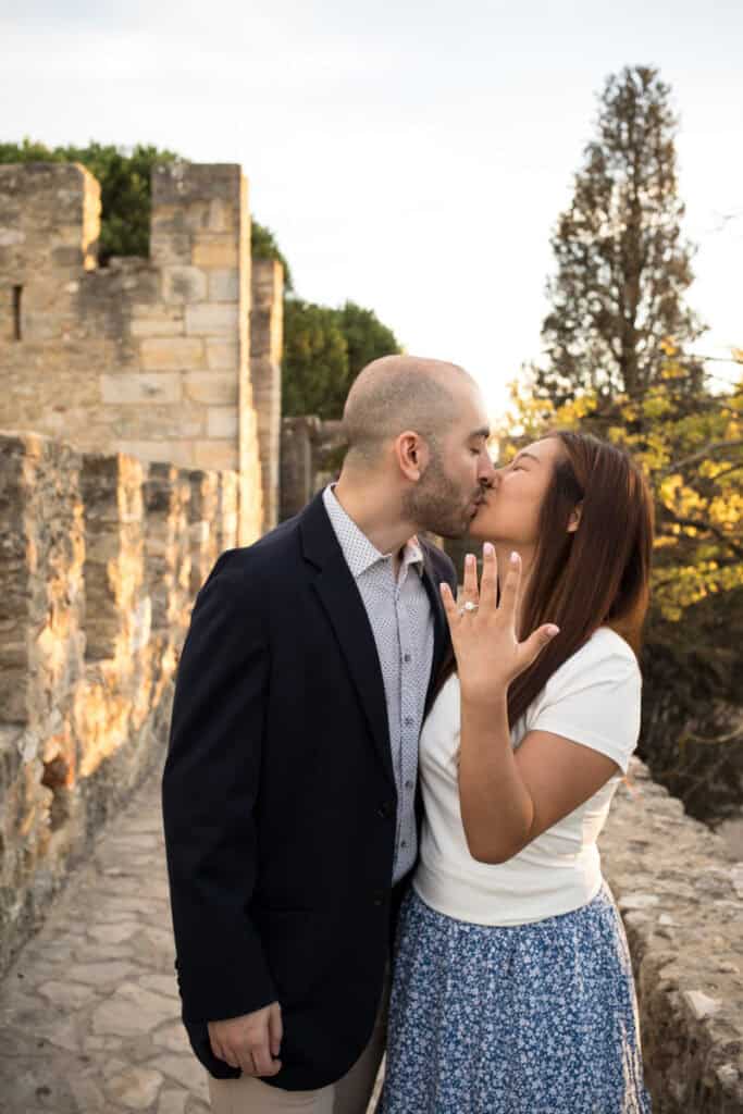 Een stralend koppel kust elkaar na een Lissabon aanzoek bij het kasteel, waarbij de vrouw haar hand met verlovingsring trots in de camera laat zien op de historische kasteelmuren bij zonsondergang.